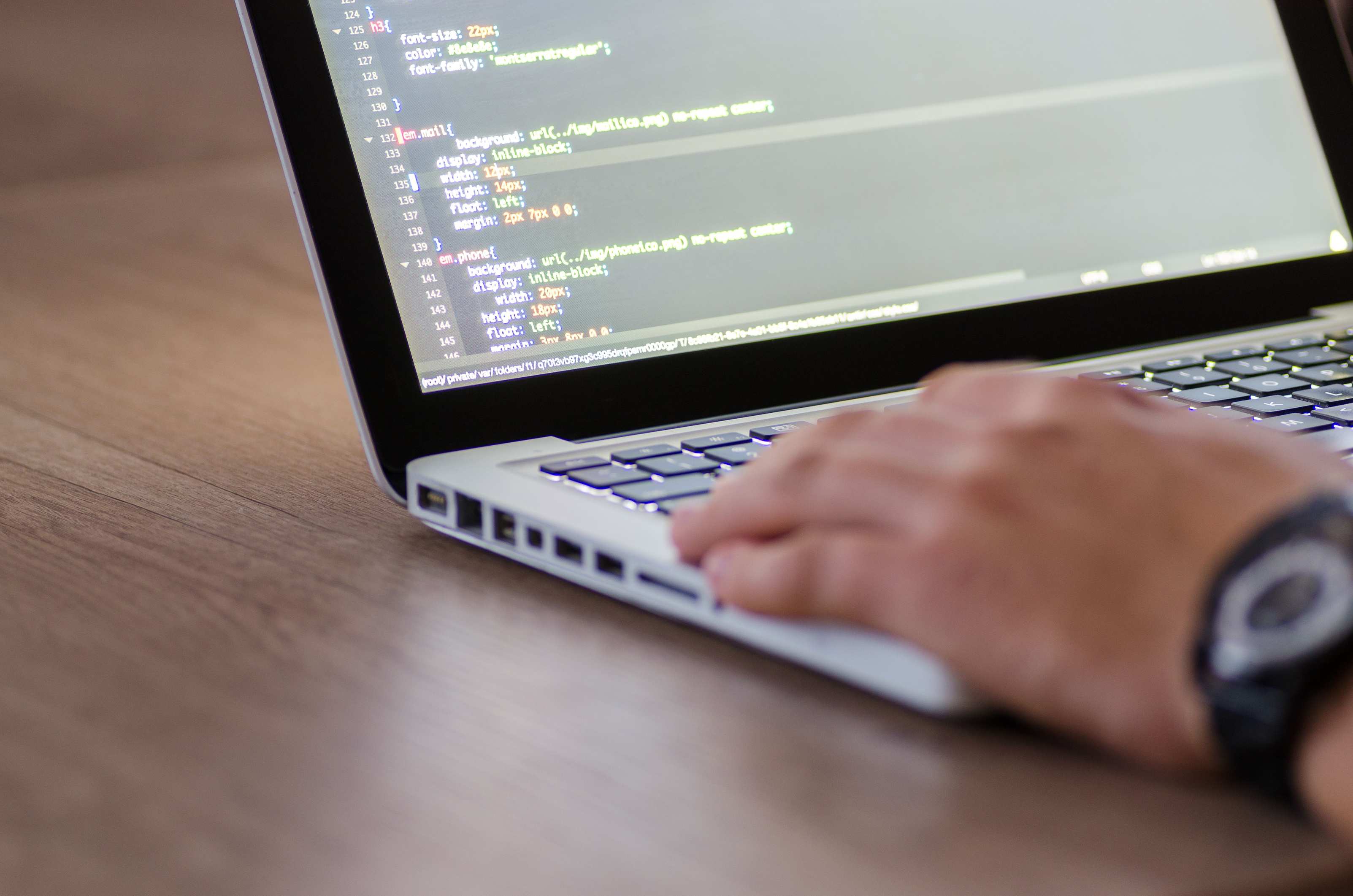 Hands typing on a keyboard beside a laptop screen, person coding on a laptop with HTML code on screen showcasing development work